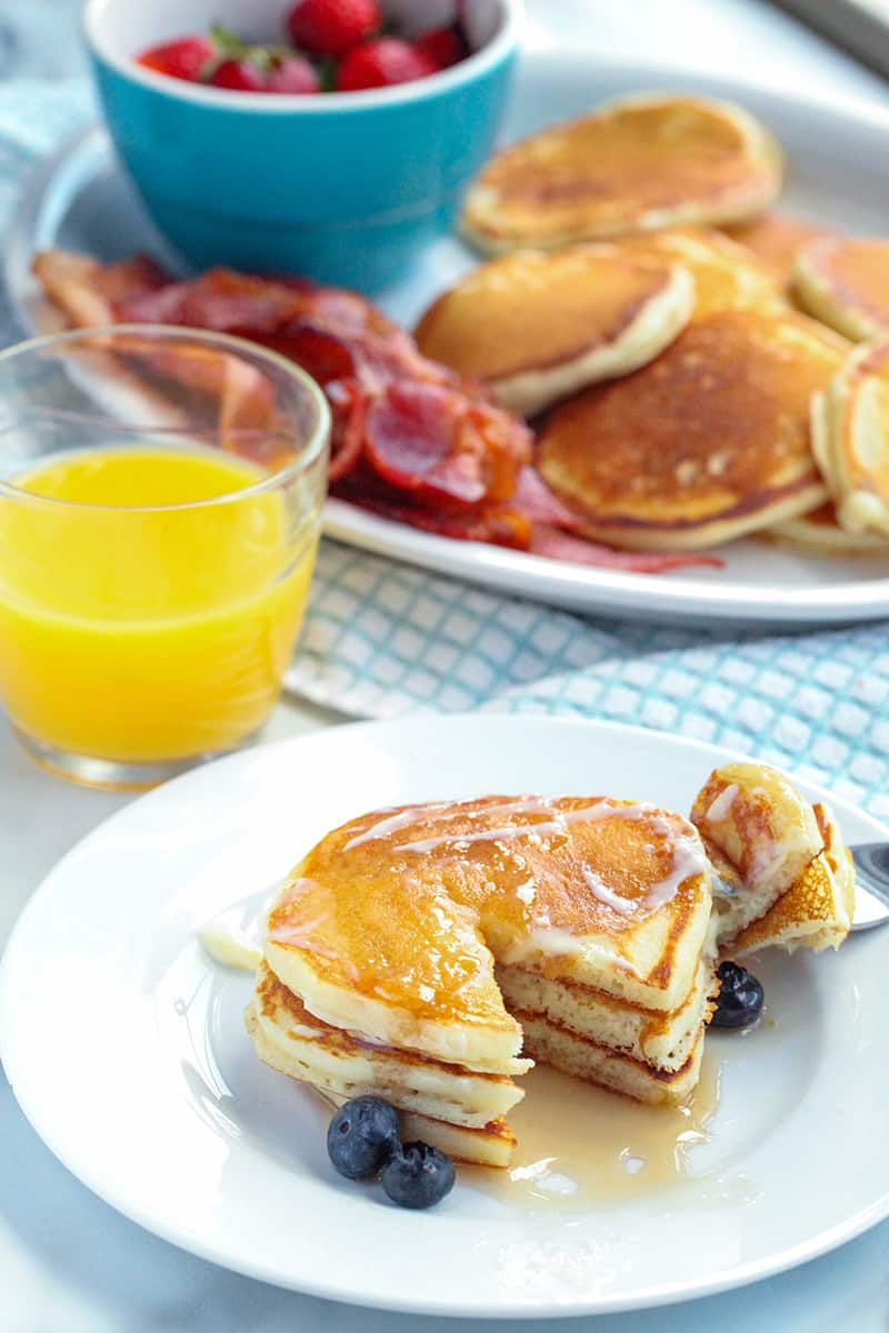 Plate with pancakes and a glass of orange juice in background with platter with bacon and pancakes in the background