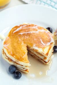 A stack of pancakes on a white plate with syrup and blueberries
