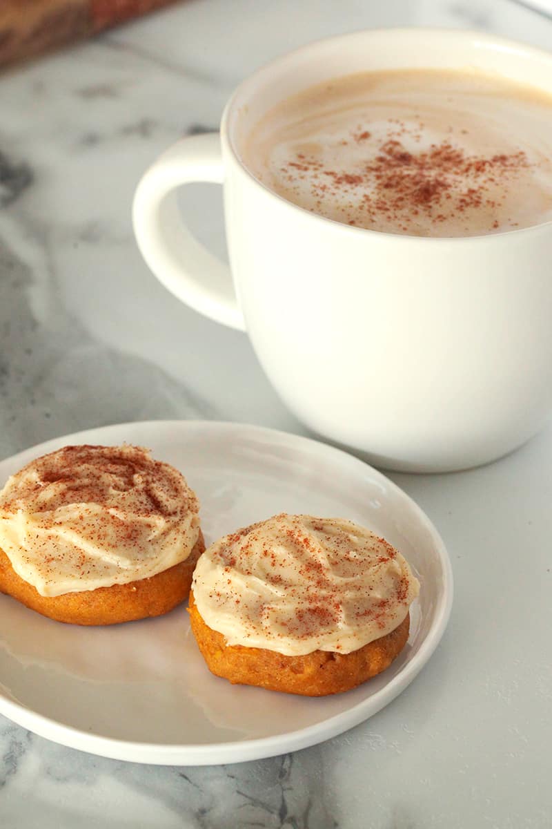 A white plate with two pumpkin cookies on to with a cup of coffee in the background